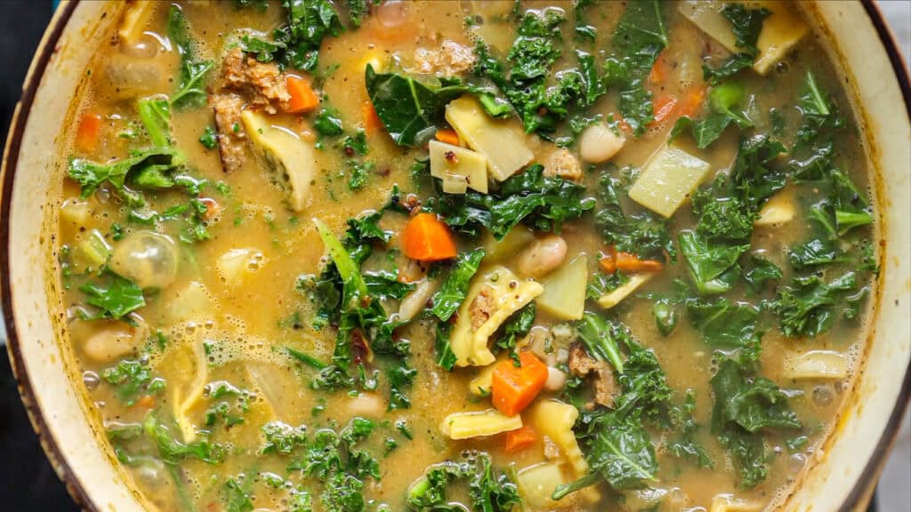 A close-up of a hearty vegetable kale white bean soup filled with leafy kale, carrots, beans, potatoes, and other vegetables in a light broth, simmering in a pot.